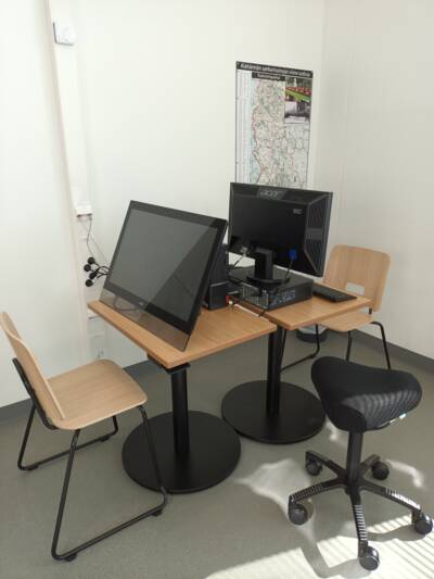 Kaksi tietokonepöytää vastakkain kirjastossa. Toisella pöydällä on lehtienlukulaite ja toisella pöytätietokone.