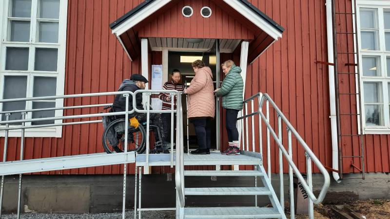 punainen rakennus, jonka edustalla portaat ja ramppi, ovella ihmisiä, joista yksi pyörätuolissa.